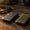 Two portable SSDs on a workbench in warm light, one smooth charcoal and one textured olive with a metal attachment loop, beside a USB-C cable, a lens cap and a camera bag